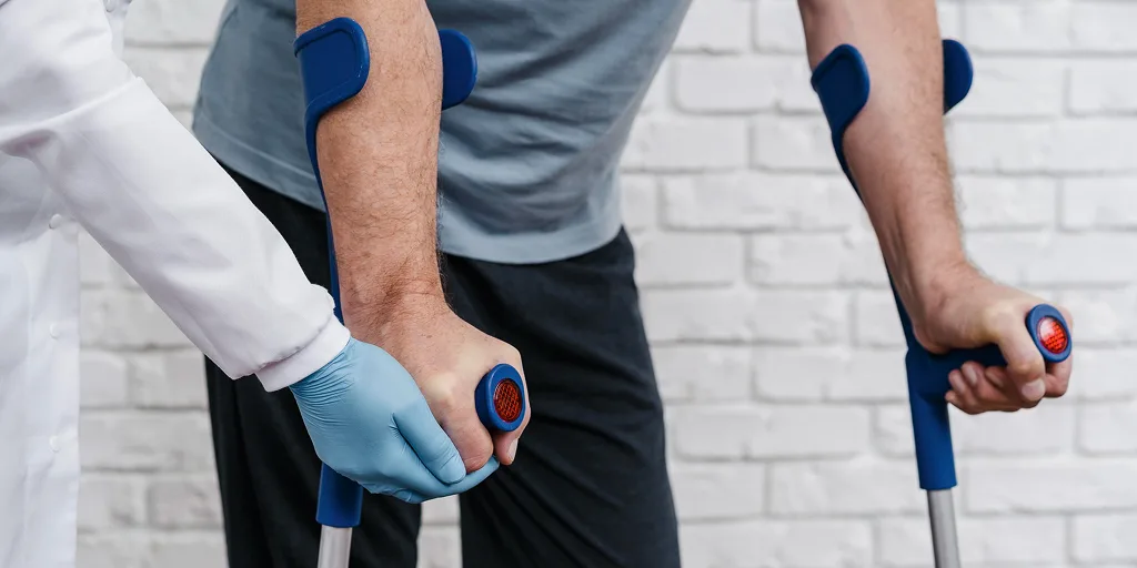 Close-up of a patient using forearm crutches, assisted by a healthcare professional wearing gloves. Image represents mobility support after an ACL injury.