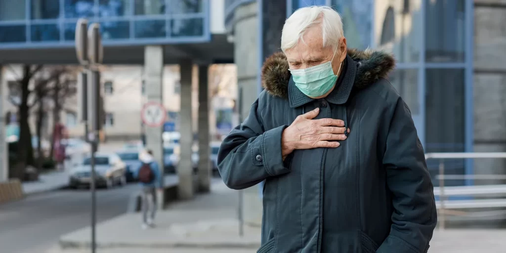 Older man wearing a mask holding chest, showing symptoms of heart pain or heart attack on a cold winter day