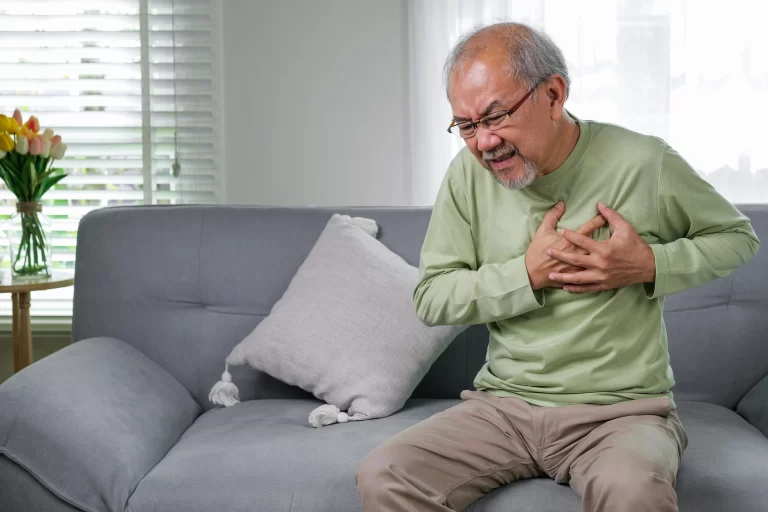 Elderly man clutching his chest in pain while sitting on a sofa, showing signs of possible heart attack or severe chest discomfort.