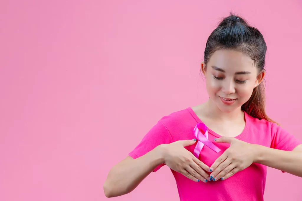 A woman wearing a pink shirt with a pink ribbon symbolizing breast cancer awareness, gently placing her hands on her chest against a pink background.