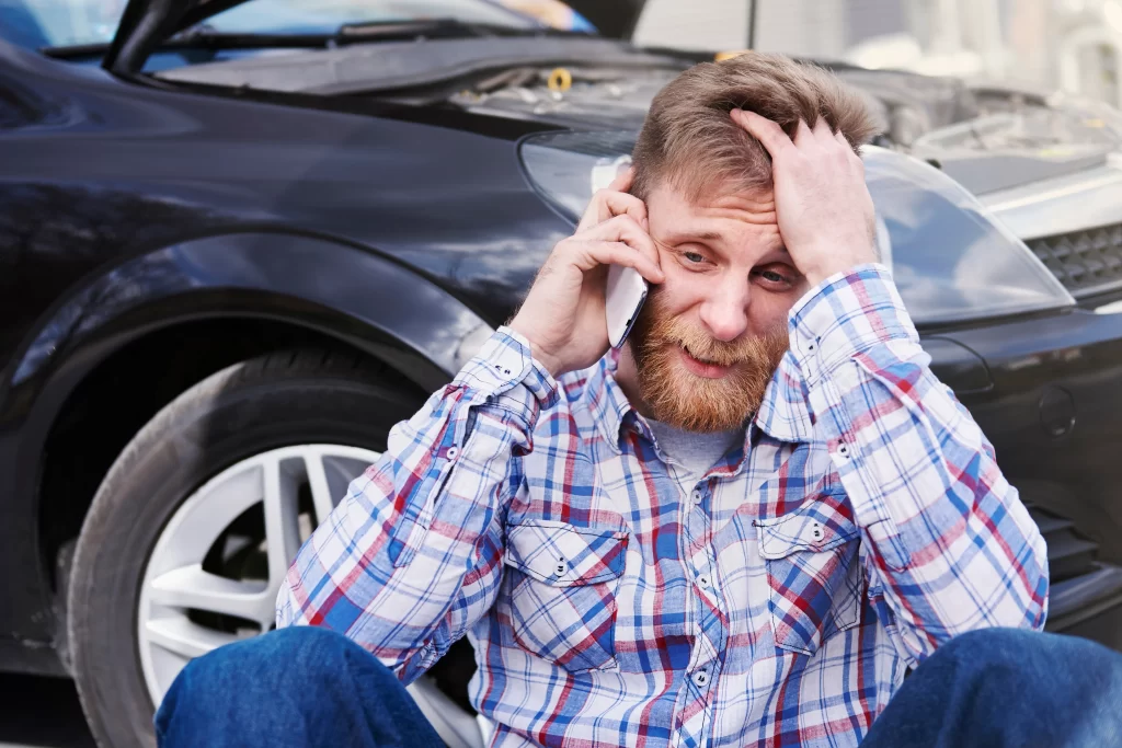Stressed man calling for help after a car accident with damaged vehicle in background