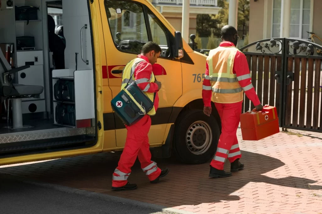 Two paramedics wearing red uniforms and yellow reflective vests are walking toward a building, carrying medical equipment bags. A yellow ambulance with its side door open is parked nearby, showing the interior with emergency medical gear.