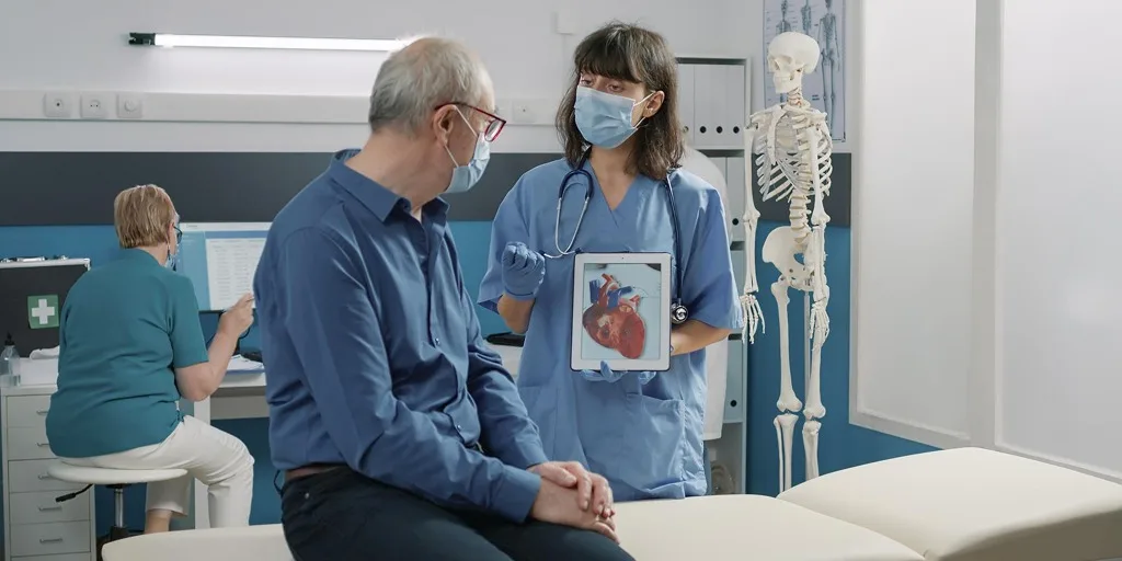 Female healthcare professional wearing a mask showing a digital heart illustration to an elderly male patient during a consultation in a medical clinic.