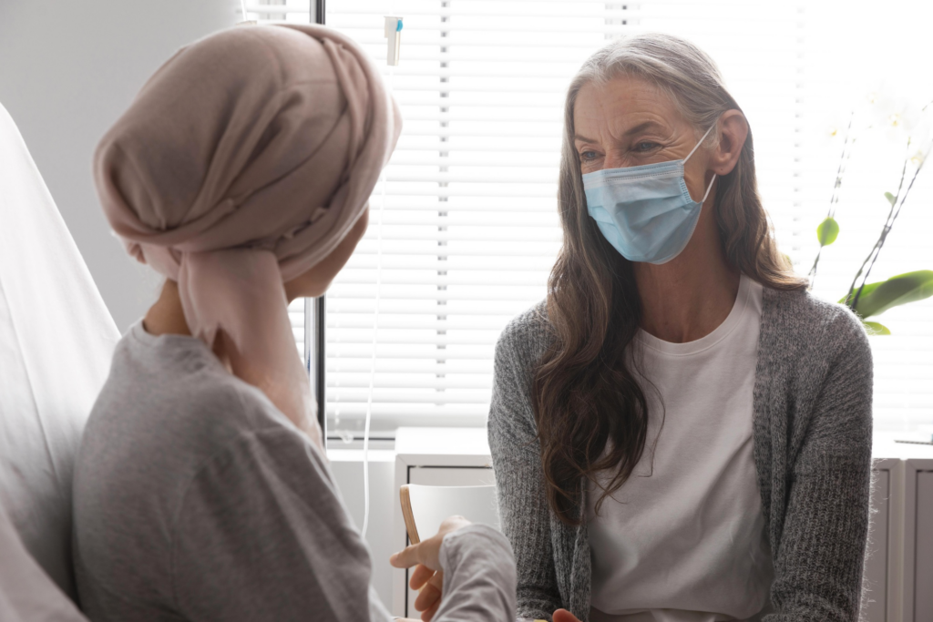 A woman wearing a headscarf sits in a hospital room talking with another woman wearing a medical mask. Both appear engaged in a supportive conversation in a clinical setting.