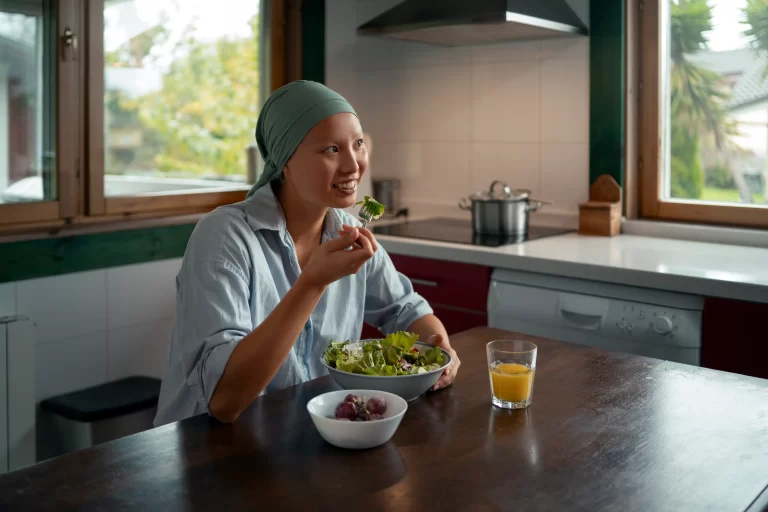 A woman wearing a headscarf sits at a kitchen table, eating a fresh green salad with grapes and a glass of juice beside her.