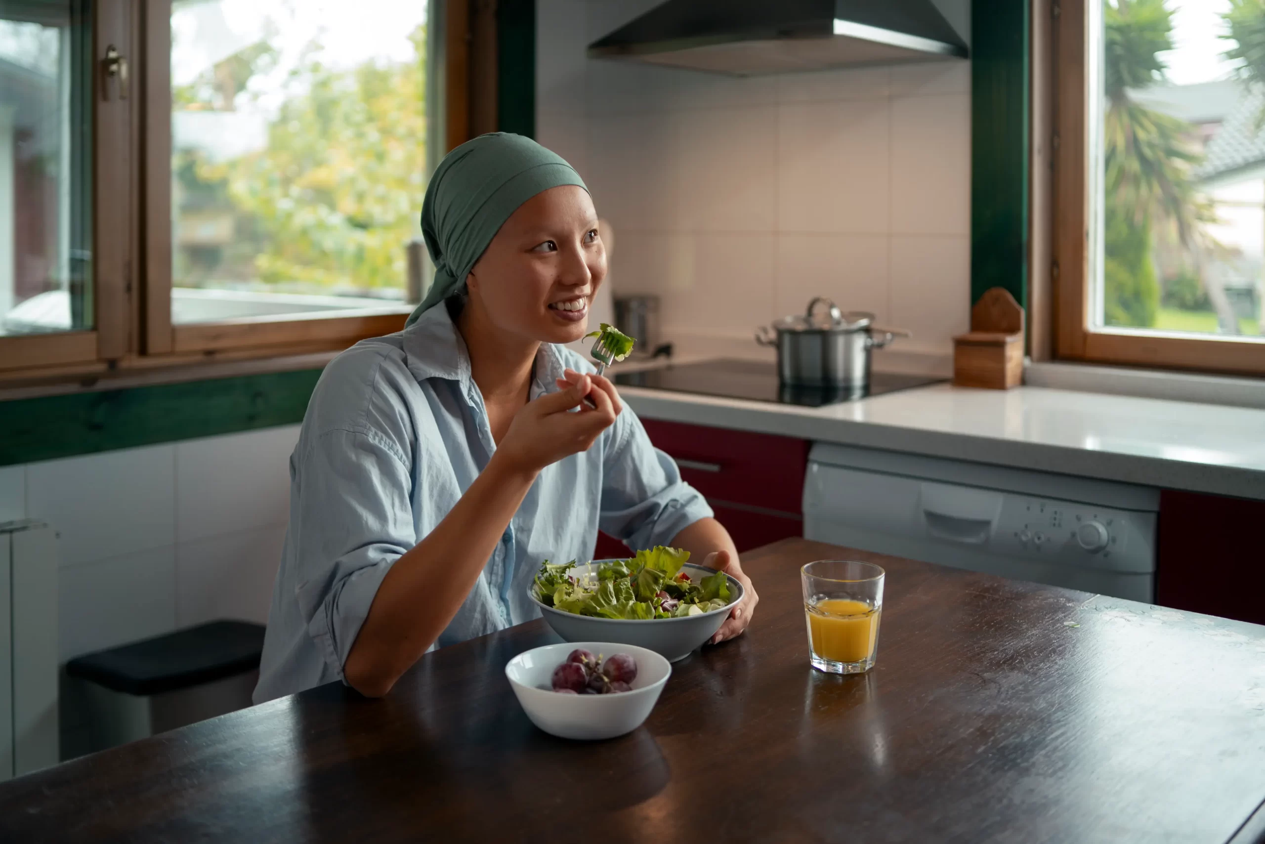 A woman wearing a headscarf sits at a kitchen table, eating a fresh green salad with grapes and a glass of juice beside her.